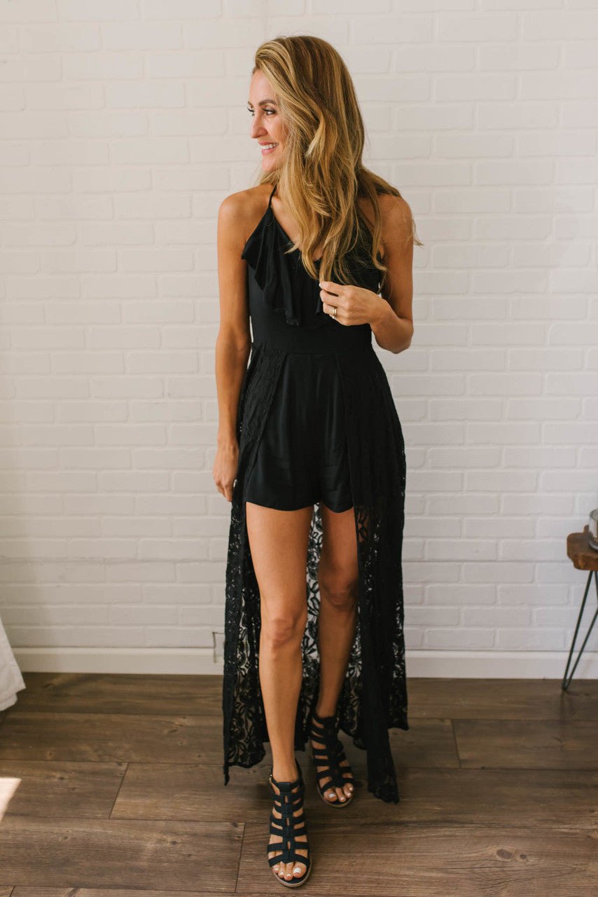 Woman in the Kiss Me Goodnight Maxi Romper - Black, standing on a wooden floor against a white brick wall.