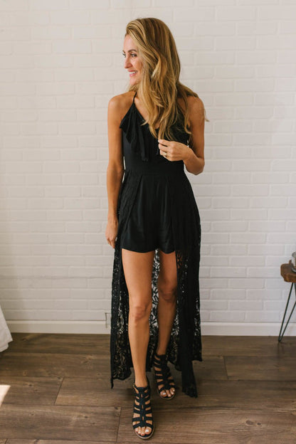 Woman in the Kiss Me Goodnight Maxi Romper - Black, standing on a wooden floor against a white brick wall.
