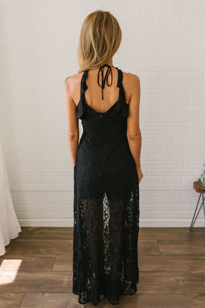 Woman with long blonde hair wears the Kiss Me Goodnight Maxi Romper - Black, posing with her back to the camera in a bright room.