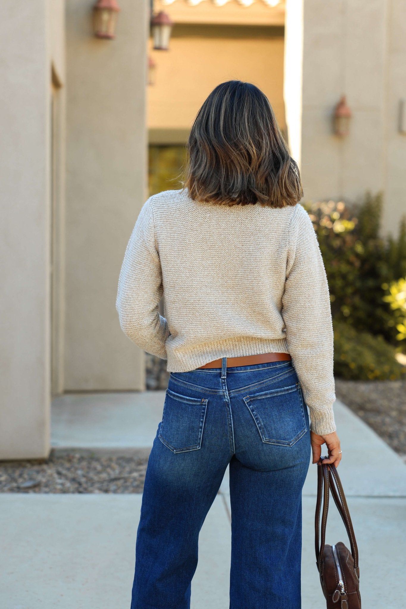 A woman with shoulder-length hair stands outside, facing away, wearing the Kylie Mocha Cable Knit Sweater and blue jeans.