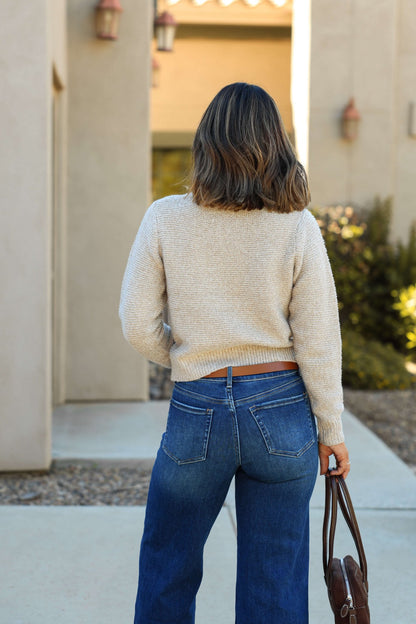 A woman with shoulder-length hair stands outside, facing away, wearing the Kylie Mocha Cable Knit Sweater and blue jeans.