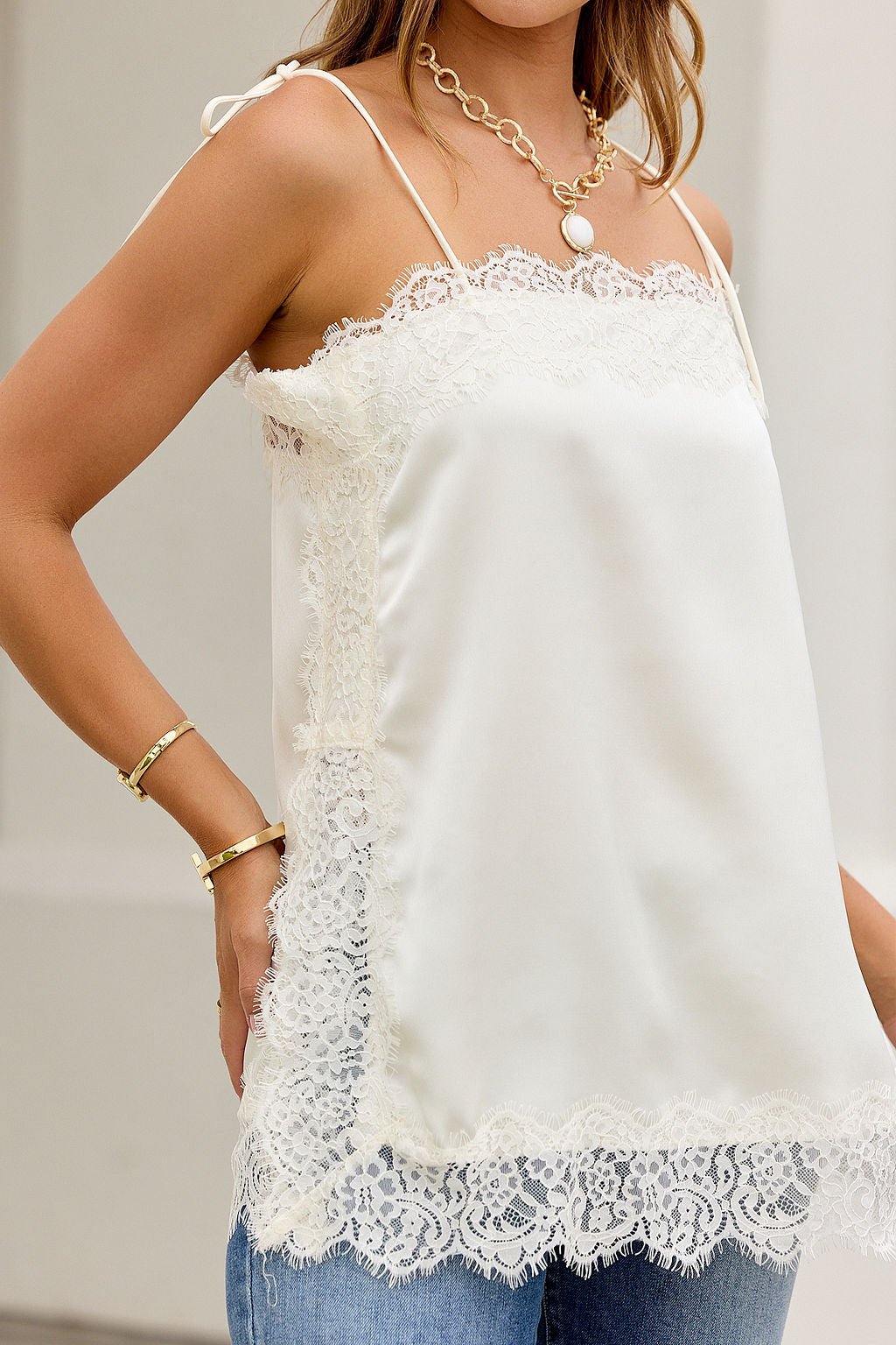 Woman in Lace Dream Cami Tank Top - Ivory with gold jewelry, blue jeans, and adjustable tie straps against a neutral background.