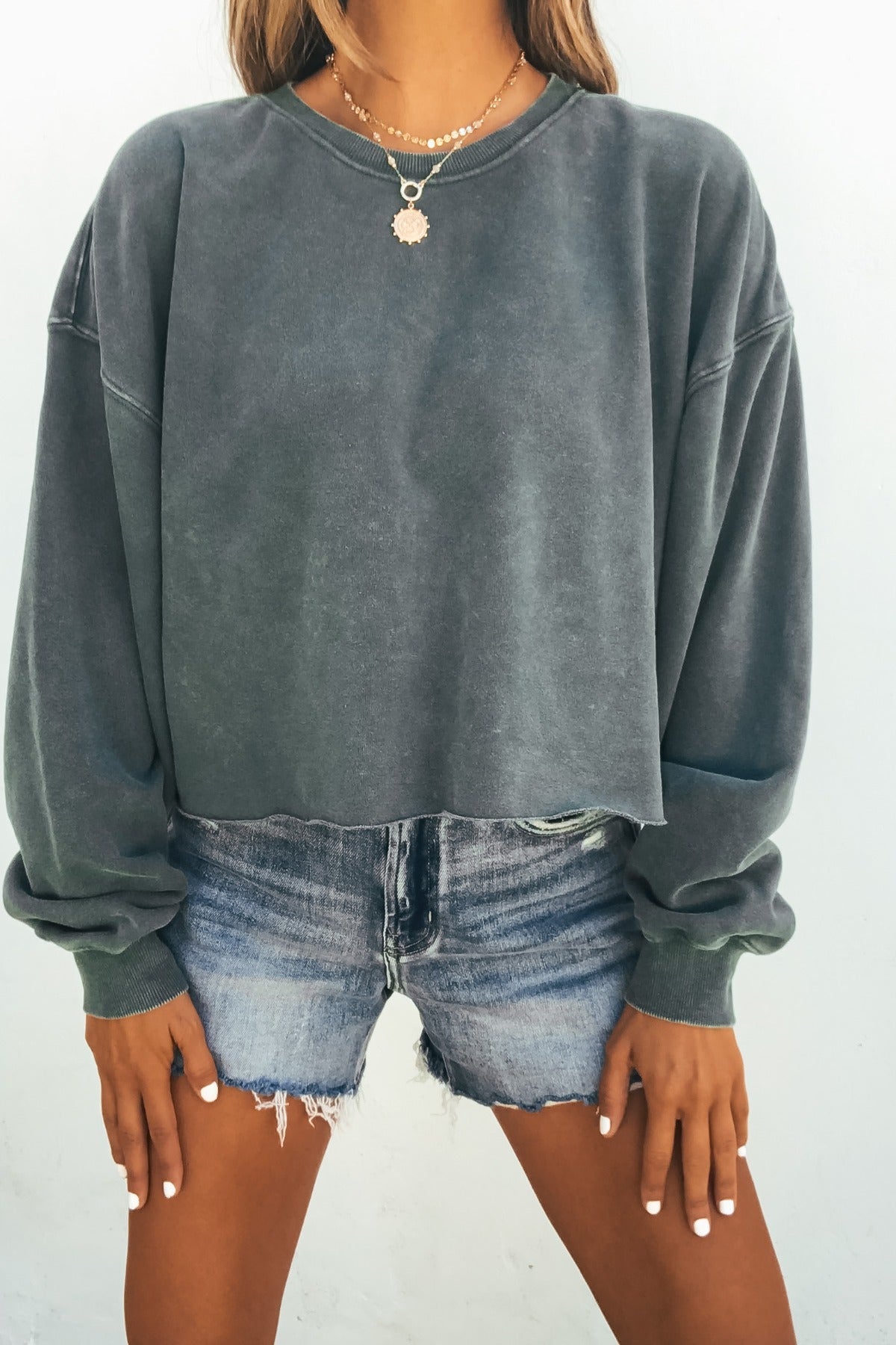 Woman in Laid-Back Crewneck Pullover - Dark Green and denim shorts, layered necklaces, stands against a light background.