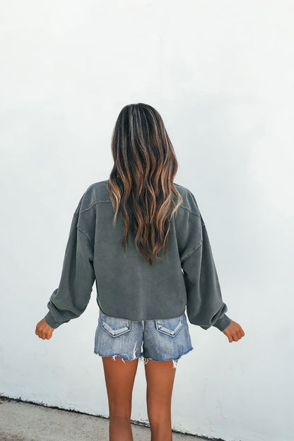Person with long wavy hair wears a relaxed fit dark green Laid-Back Crewneck Pullover and denim shorts, back to camera, white wall.