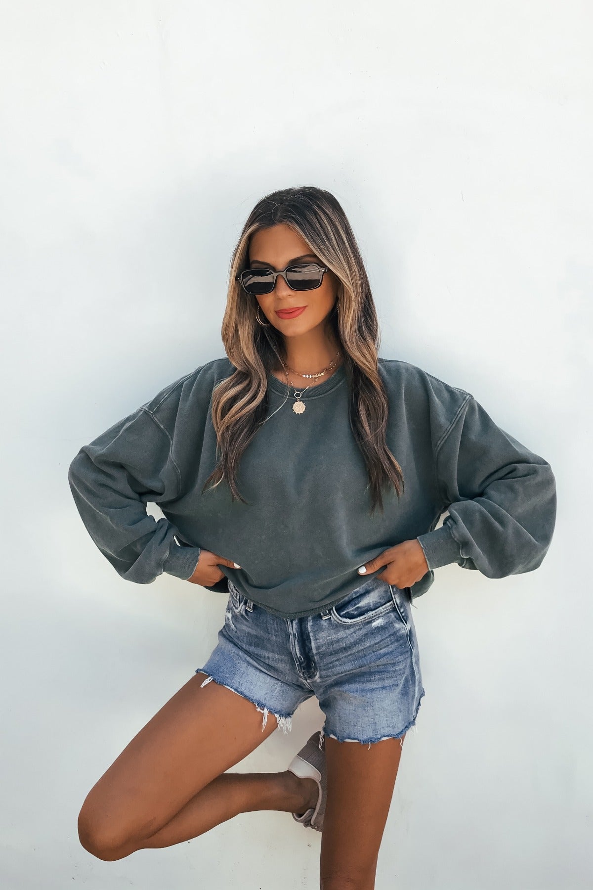 Woman in sunglasses models the Laid-Back Crewneck Pullover in dark green with denim shorts, posing confidently against a white wall.
