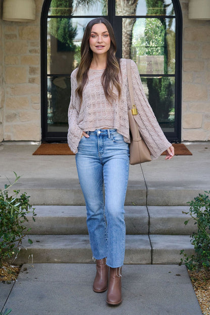 A woman stands on stone steps in a Laurel Taupe Open Knit Sweater, blue jeans, brown boots, and a beige shoulder bag by a glass door.