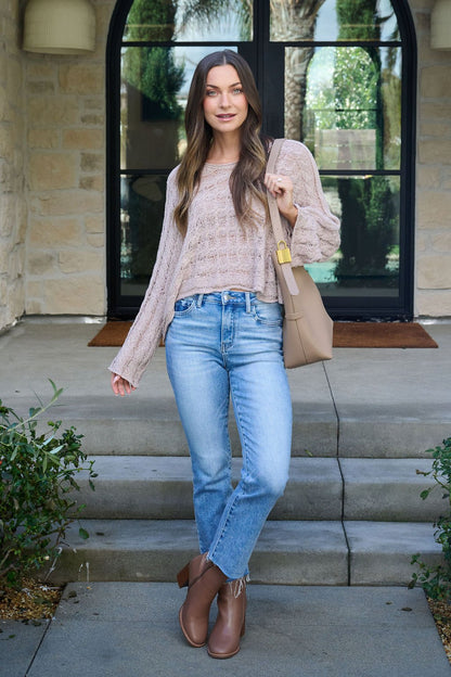 Wearing the Laurel Taupe Open Knit Sweater, a woman in jeans and brown boots smiles on steps outside a house with a beige handbag.