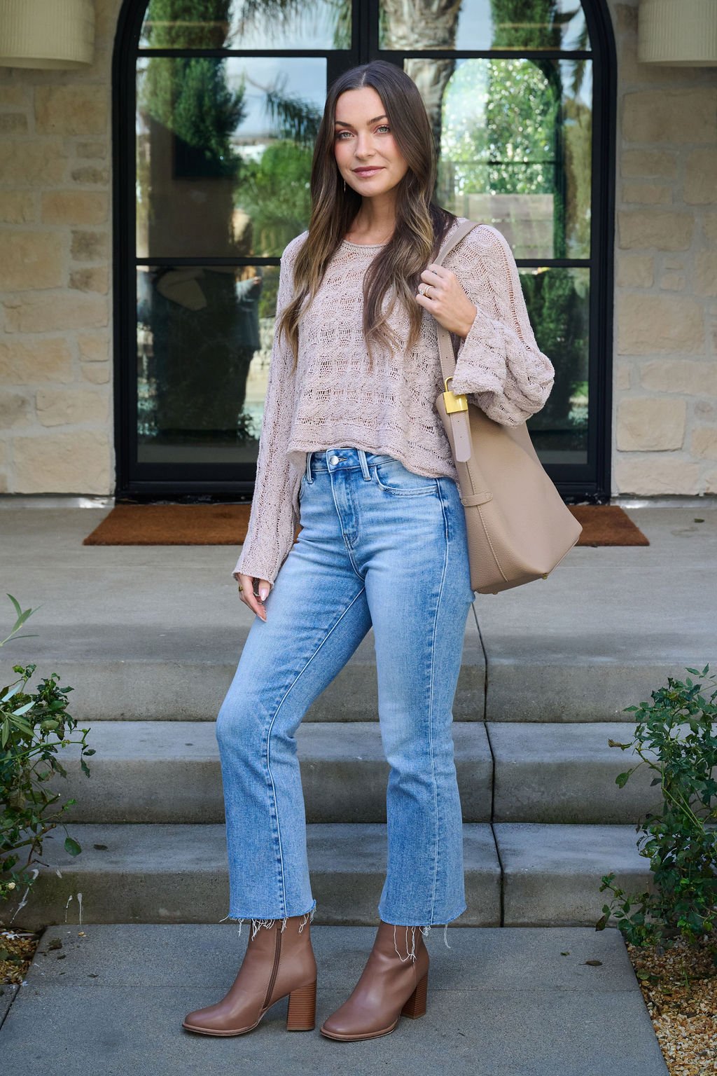 A woman stands on stone steps wearing the Laurel Taupe Open Knit Sweater, light blue jeans, tan ankle boots, and a beige tote bag.