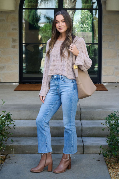 A woman stands on stone steps wearing the Laurel Taupe Open Knit Sweater, light blue jeans, tan ankle boots, and a beige tote bag.