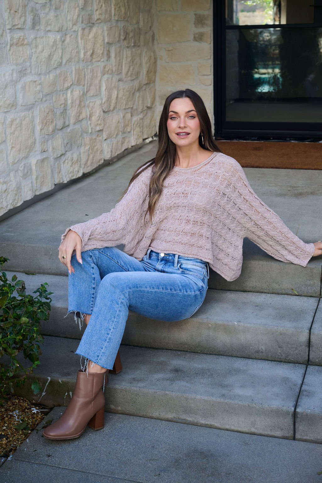 Woman with long brown hair wears the Laurel Taupe Open Knit Sweater, blue jeans, and brown ankle boots, sitting on outdoor steps.
