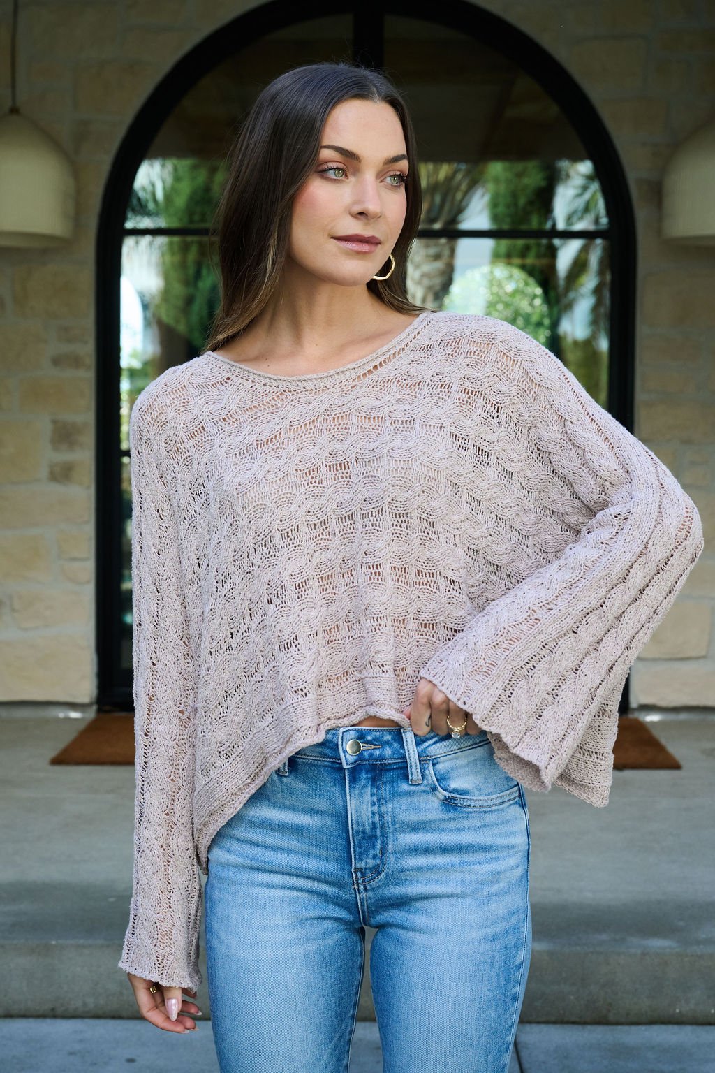Woman in the Laurel Taupe Open Knit Sweater and blue jeans stands outside by an arched doorway, looking to the side.