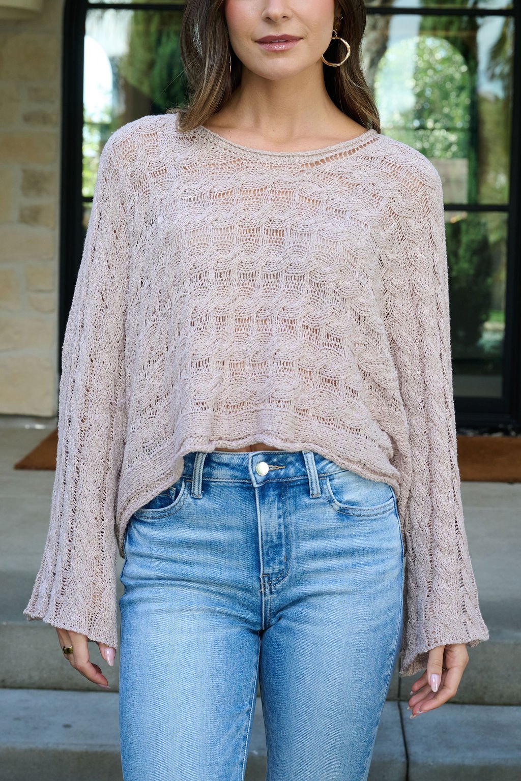 A woman in the Laurel Taupe Open Knit Sweater and light blue high-waisted jeans stands outdoors in front of a building.