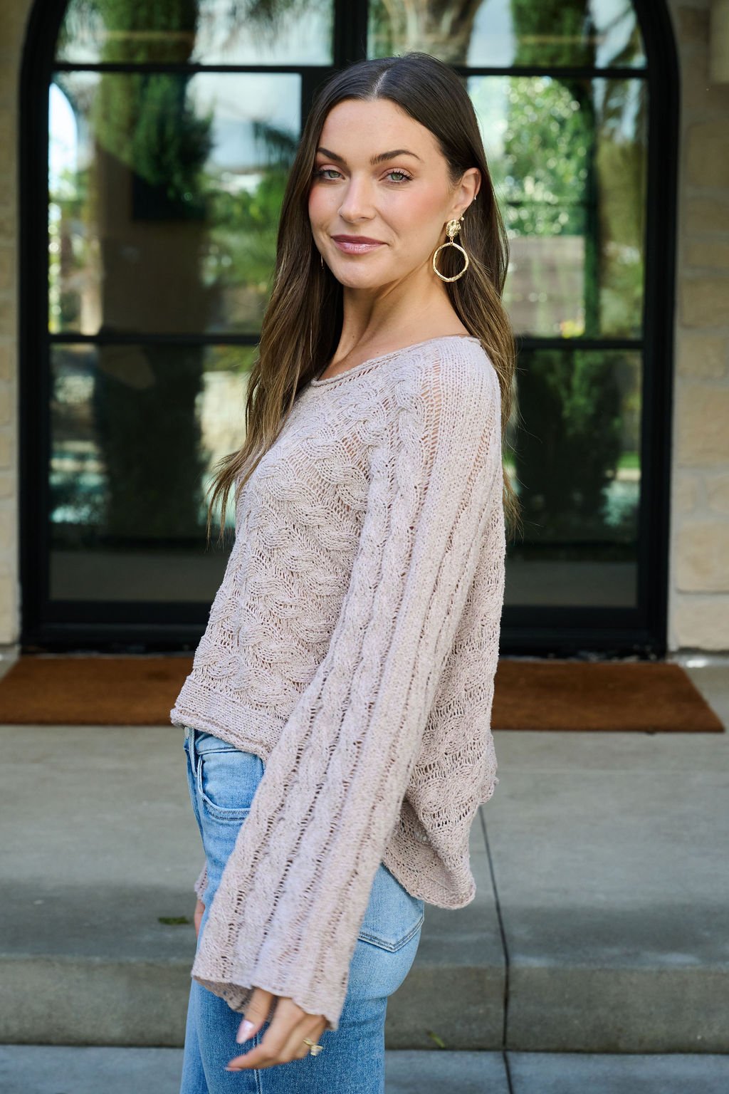 Woman with long brown hair in the Laurel Taupe Open Knit Sweater and jeans smiles outside in front of glass doors.