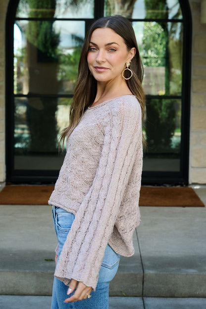 Woman with long brown hair in the Laurel Taupe Open Knit Sweater and jeans smiles outside in front of glass doors.