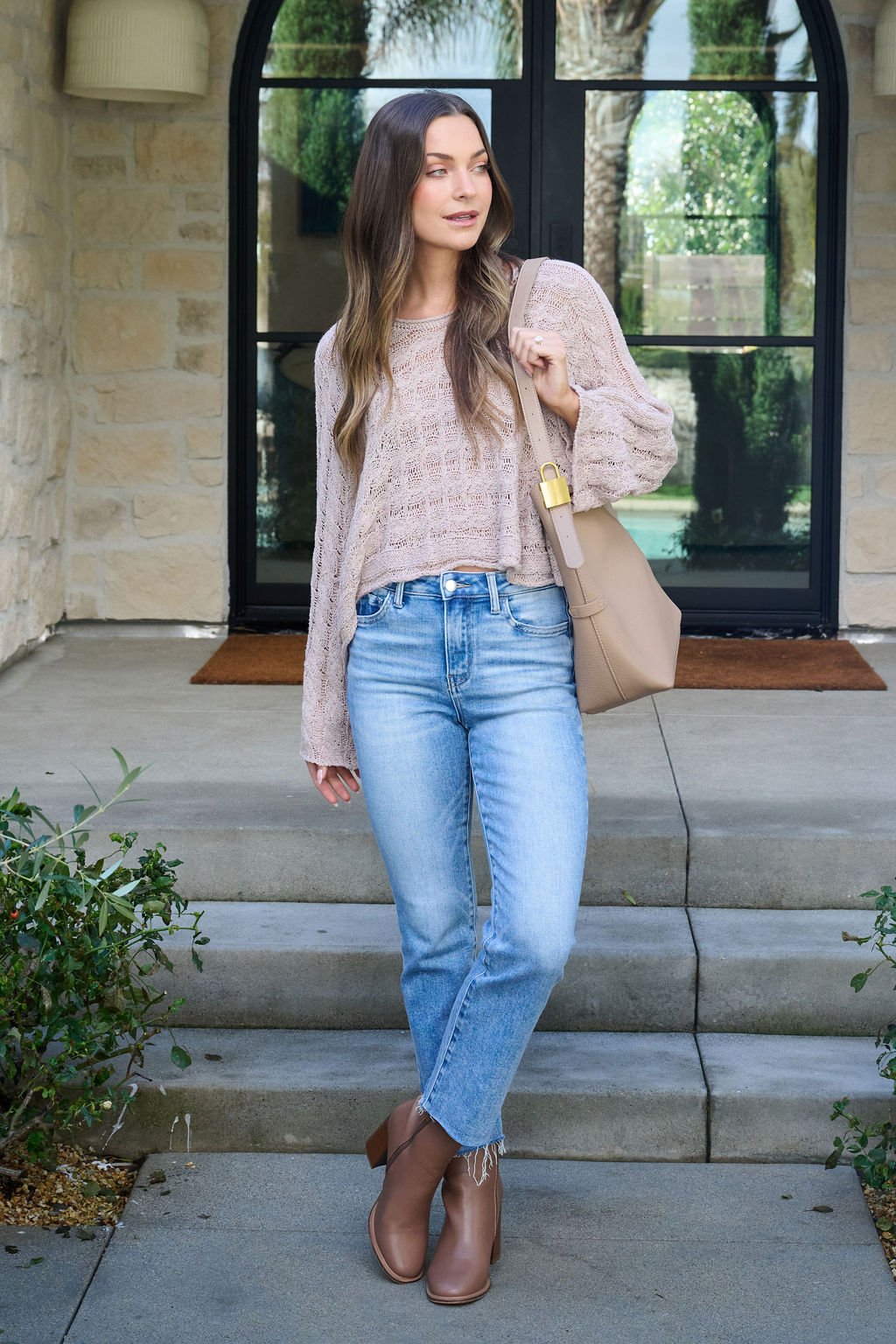 A woman in the Laurel Taupe Open Knit Sweater, blue jeans, and brown boots stands outdoors holding a beige handbag.