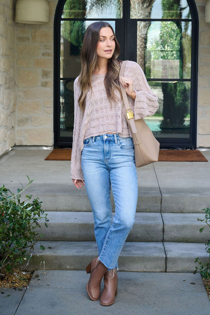 A woman in the Laurel Taupe Open Knit Sweater, blue jeans, and brown boots stands outdoors holding a beige handbag.