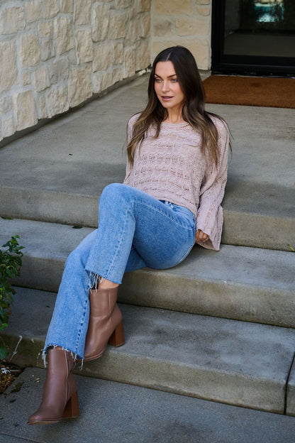 A woman in a Laurel Taupe Open Knit Sweater sits relaxed on outdoor steps beside a stone wall.