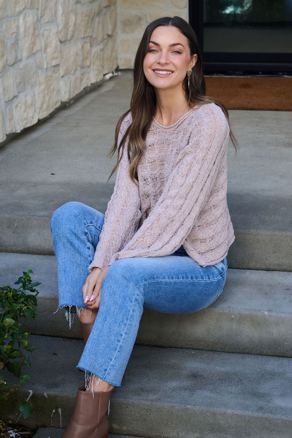 A woman smiles on outdoor steps wearing the Laurel Taupe Open Knit Sweater, blue jeans, and brown boots.