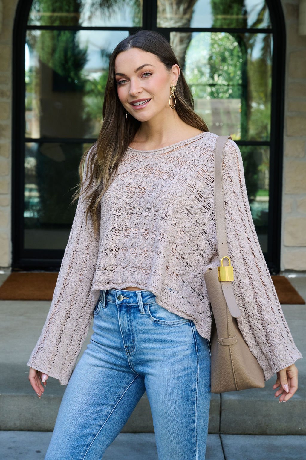 A woman in a Laurel Taupe Open Knit Sweater and blue jeans stands outside, smiling with a beige shoulder bag and gold earrings.