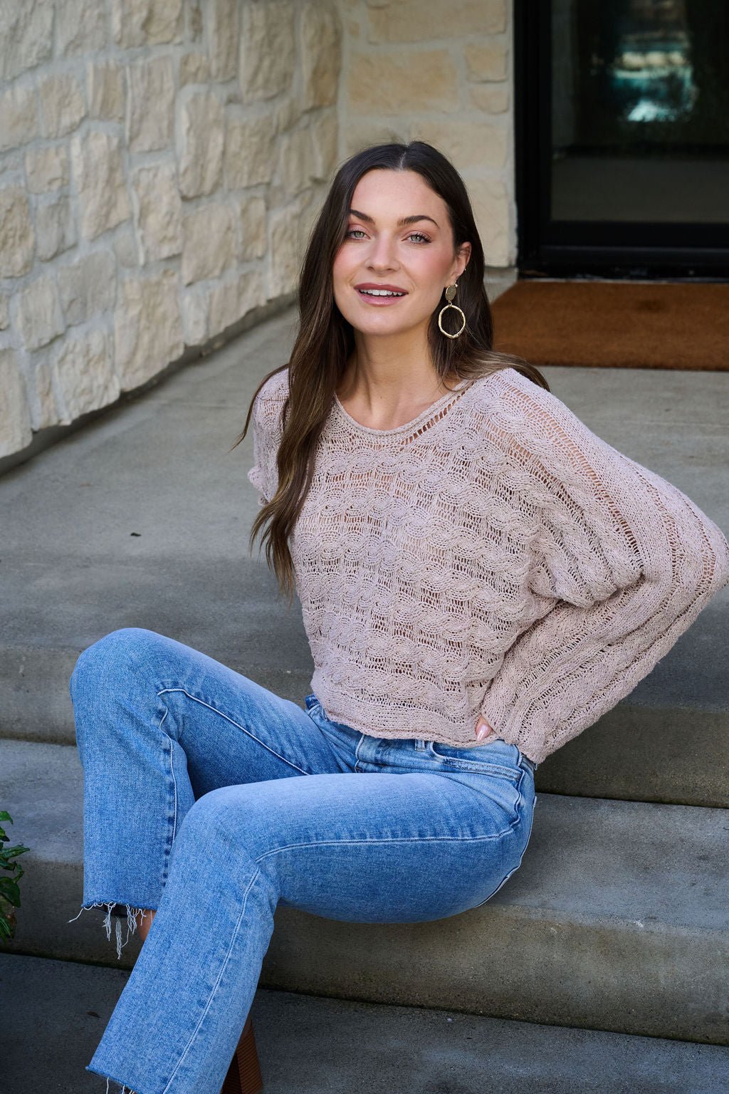 Woman with long brown hair wears the Laurel Taupe Open Knit Sweater and jeans, smiling while seated on outdoor stone steps.