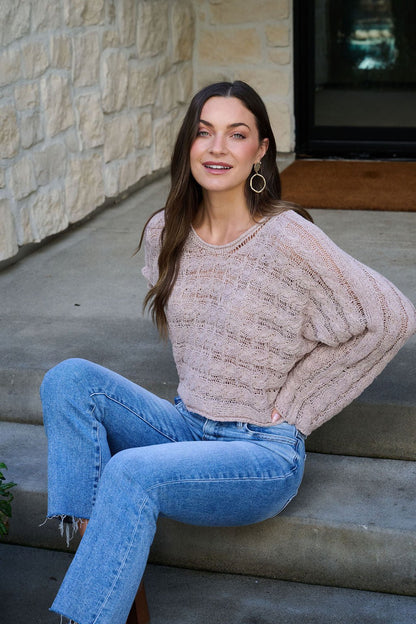 Woman with long brown hair wears the Laurel Taupe Open Knit Sweater and jeans, smiling while seated on outdoor stone steps.
