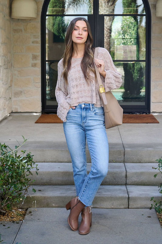 Woman stands on steps in front of a door, wearing the Laurel Taupe Open Knit Sweater - FINAL SALE, blue jeans, and brown boots.