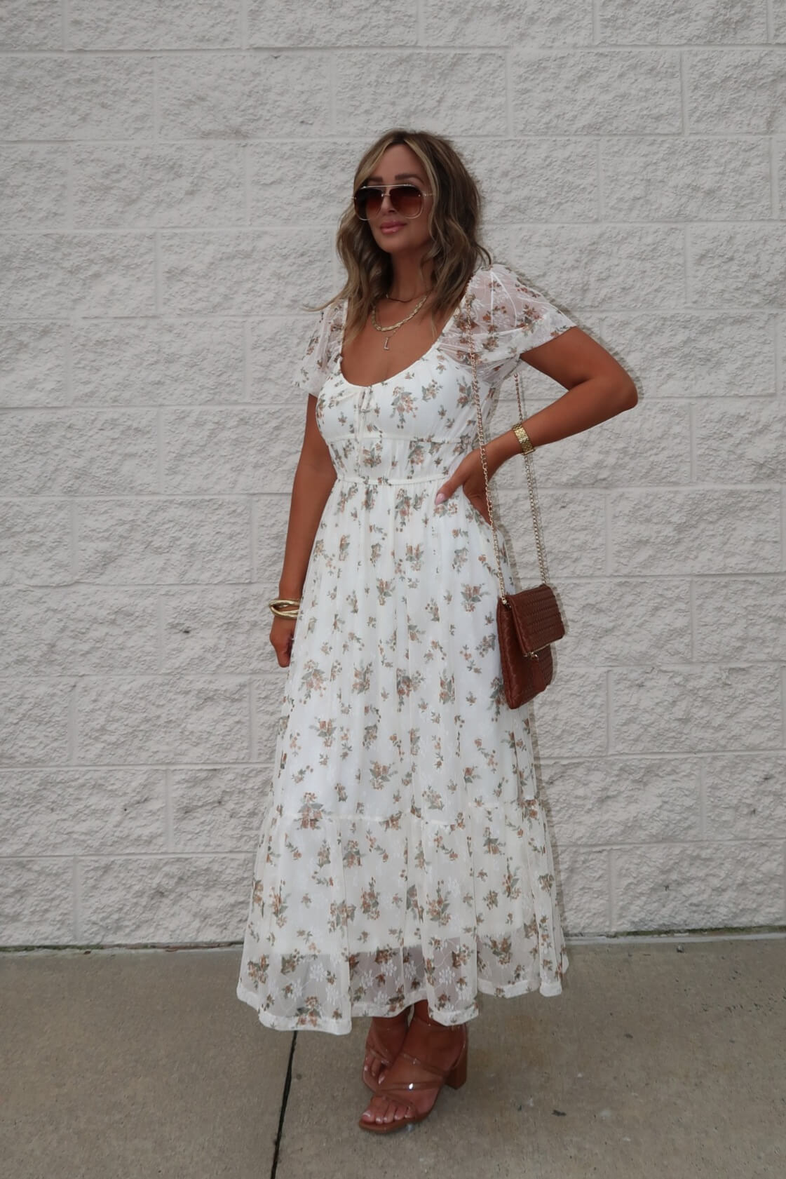 Woman in a Lauri Cream Floral Print Sweetheart Midi Dress, brown sandals, sunglasses, and a purse by a light brick wall.