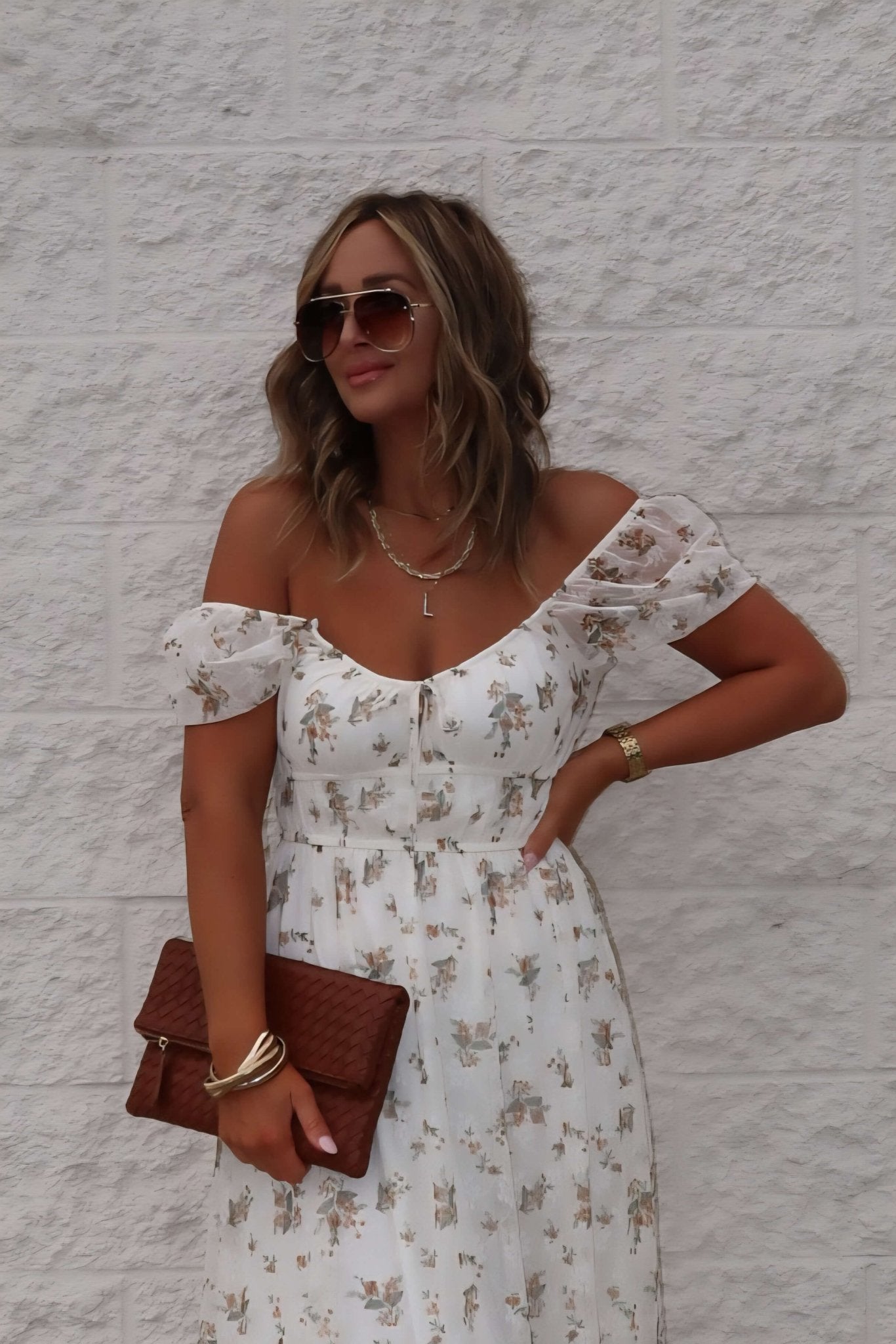 Woman wears the Lauri Cream Floral Print Sweetheart Midi Dress, holding a brown clutch and sunglasses against a white wall.