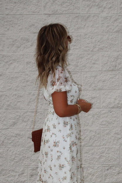 Woman in the Lauri Cream Floral Print Sweetheart Midi Dress stands sideways by a light brick wall.