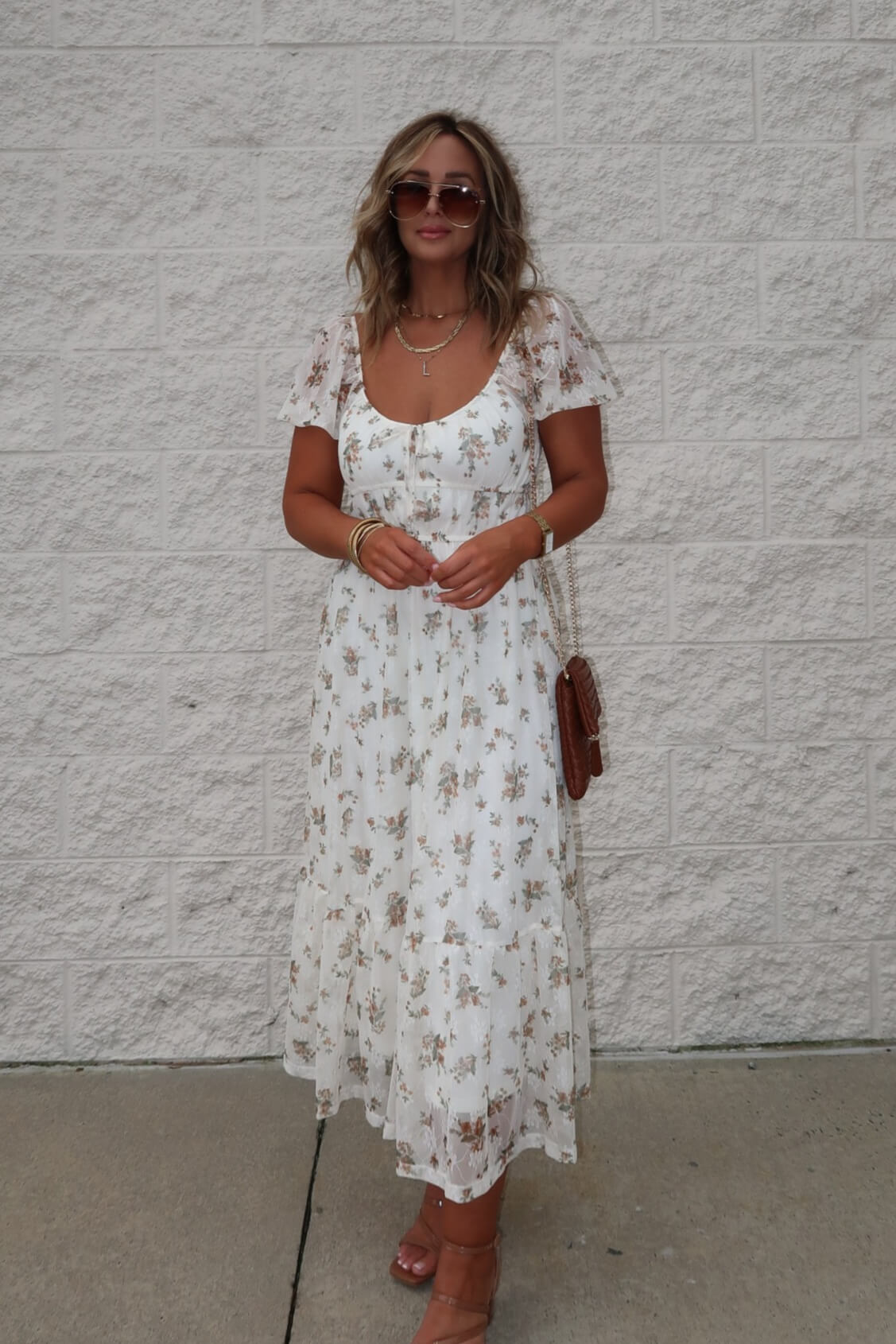 Woman in Lauri Cream Floral Print Sweetheart Midi Dress, sunglasses, sandals, holding brown purse by a light brick wall.