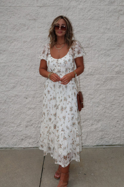 Woman in Lauri Cream Floral Print Sweetheart Midi Dress, sunglasses, sandals, holding brown purse by a light brick wall.