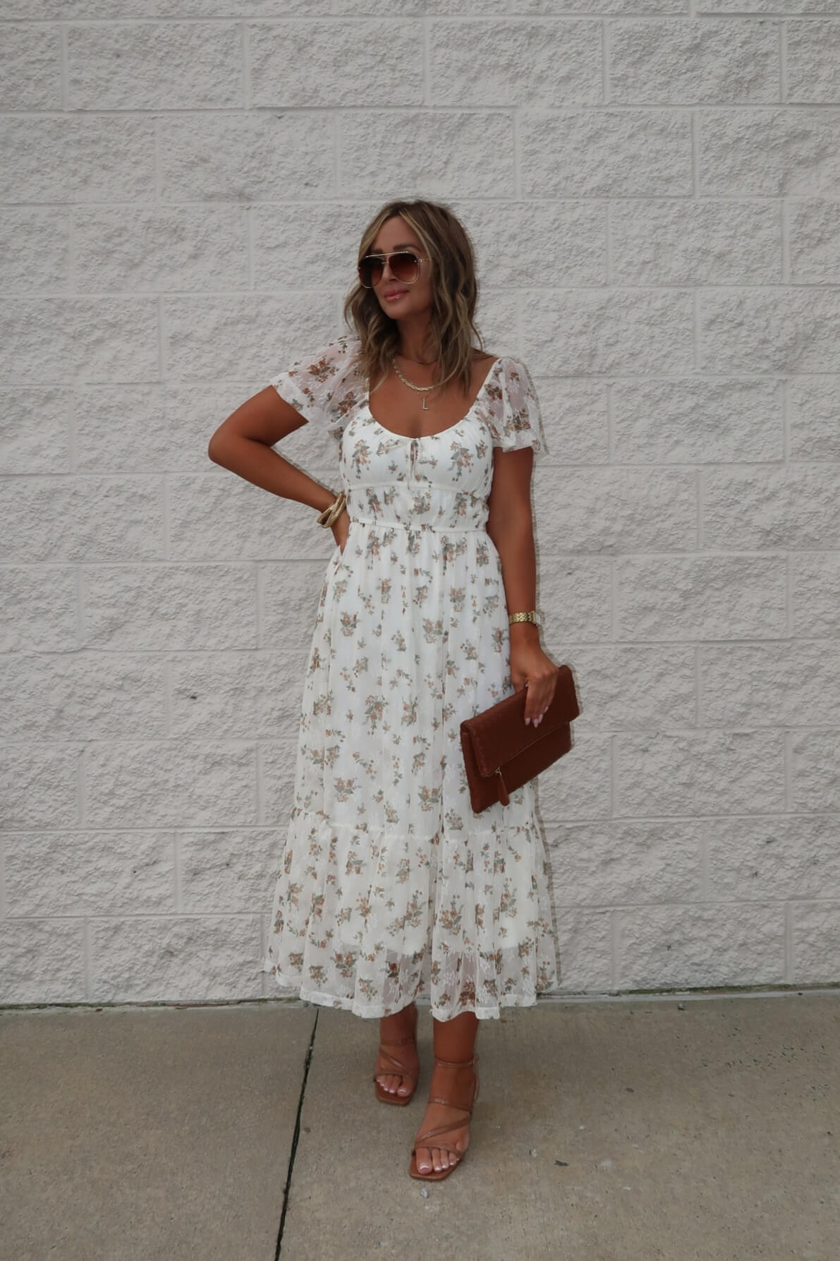 Woman in the Lauri Cream Floral Print Sweetheart Midi Dress holds a brown clutch, styled with tan sandals and sunglasses by a brick wall.
