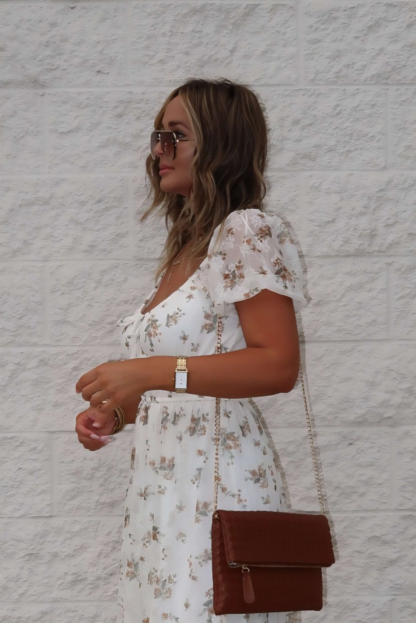 Woman wears the Lauri Cream Floral Print Sweetheart Midi Dress, sunglasses, watch, brown purse, by a white brick wall.
