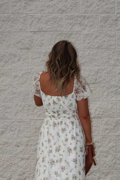 Woman with wavy hair wears the Lauri Cream Floral Print Sweetheart Midi Dress, standing by a light brick wall with a brown clutch.