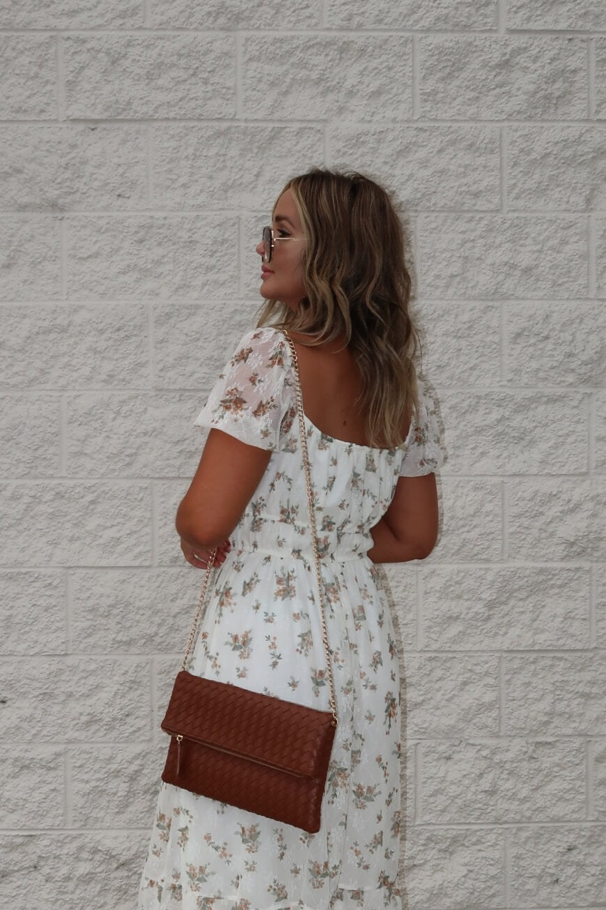 Woman in the Lauri Cream Floral Print Sweetheart Midi Dress stands with a brown purse by a textured light brick wall, looking aside.