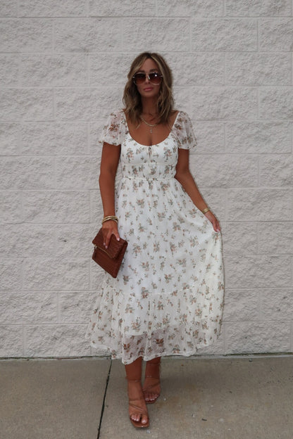 Woman in the Lauri Cream Floral Print Sweetheart Midi Dress, tan sandals, sunglasses, and brown clutch poses by a textured wall.