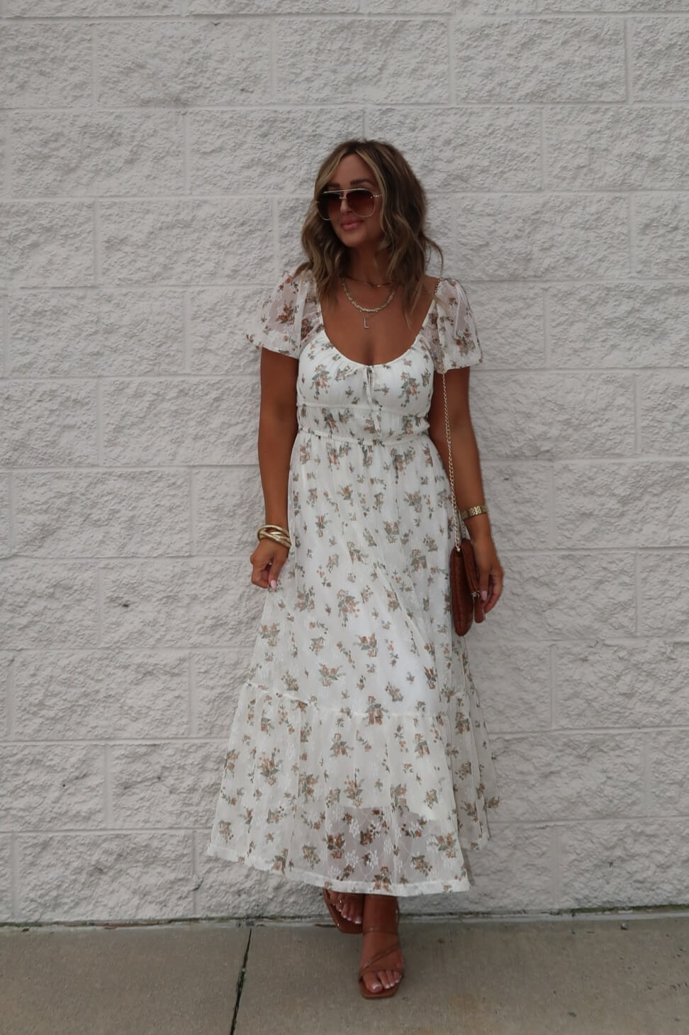 Woman in Lauri Cream Floral Print Sweetheart Midi Dress, brown sandals, sunglasses, and gold jewelry by a light brick wall.