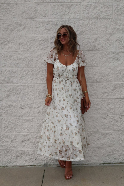 Woman in Lauri Cream Floral Print Sweetheart Midi Dress, brown sandals, sunglasses, and gold jewelry by a light brick wall.