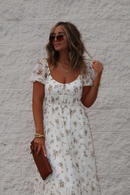 Woman in the Lauri Cream Floral Print Sweetheart Midi Dress holds a brown clutch and wears sunglasses.