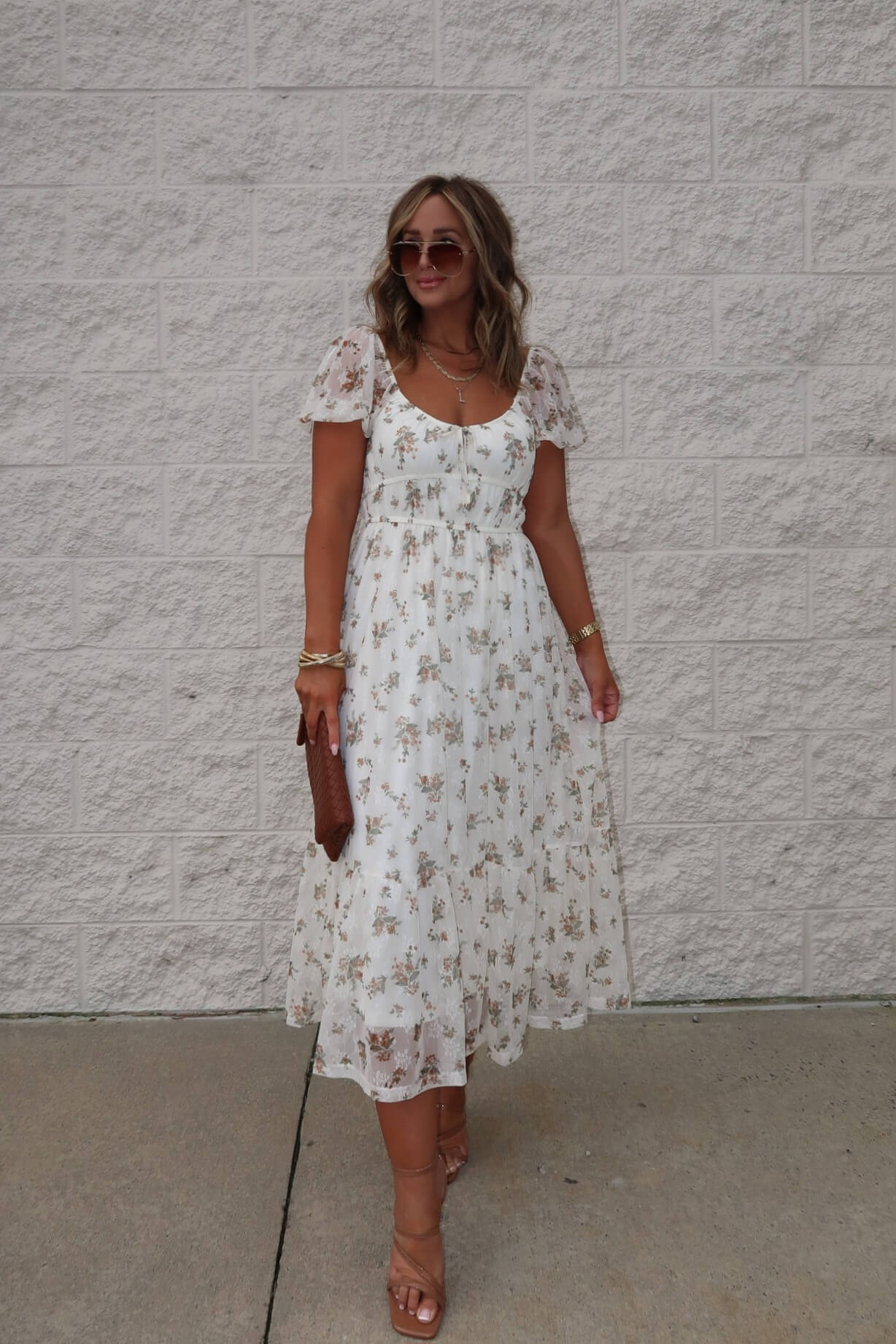 Woman in the Lauri Cream Floral Print Sweetheart Midi Dress, holding a brown clutch and wearing sunglasses by a white wall.