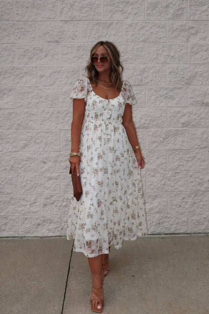 Woman in the Lauri Cream Floral Print Sweetheart Midi Dress, holding a brown clutch and wearing sunglasses by a white wall.