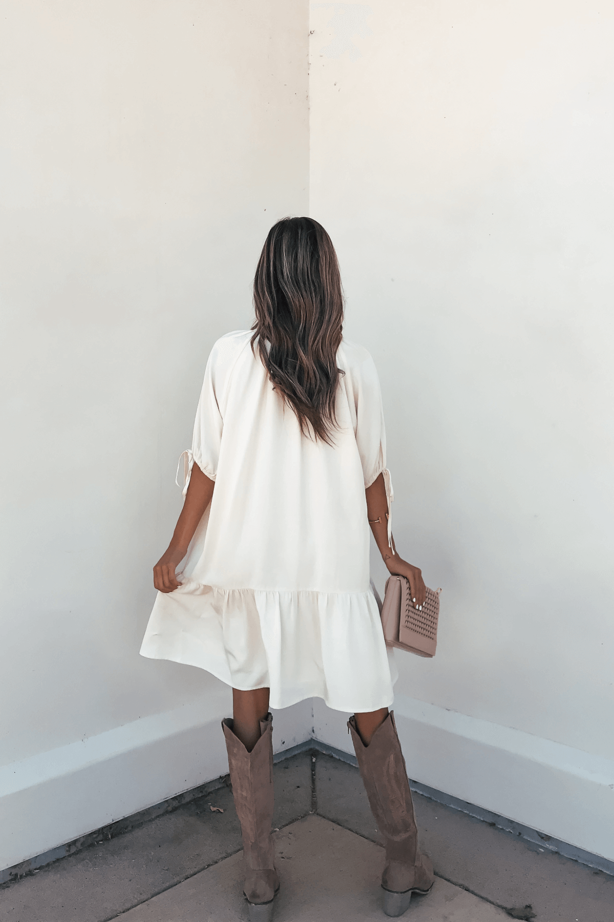 A woman in the Laurie Cream Button Down Mini Dress and brown boots holds a pink clutch while facing a white wall.