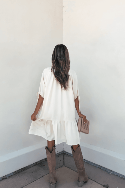 A woman in the Laurie Cream Button Down Mini Dress and brown boots holds a pink clutch while facing a white wall.