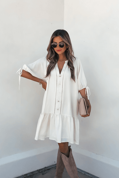 Woman in Laurie Cream Button Down Mini Dress with brown knee-high boots, sunglasses, and beige clutch by a white wall.