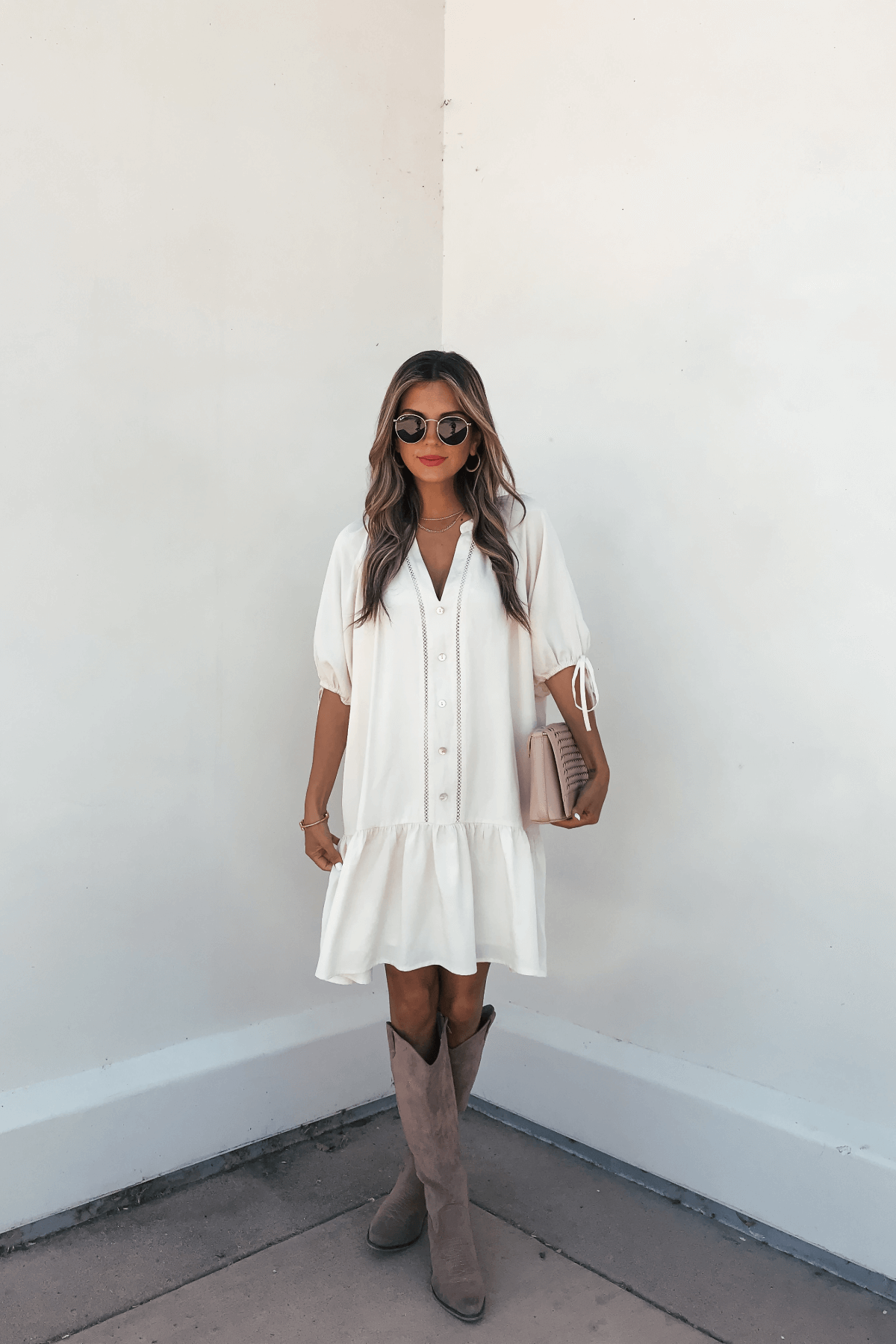 Woman in a Laurie Cream Button Down Mini Dress, taupe knee-high boots, sunglasses, and pink bag stands by a white wall.