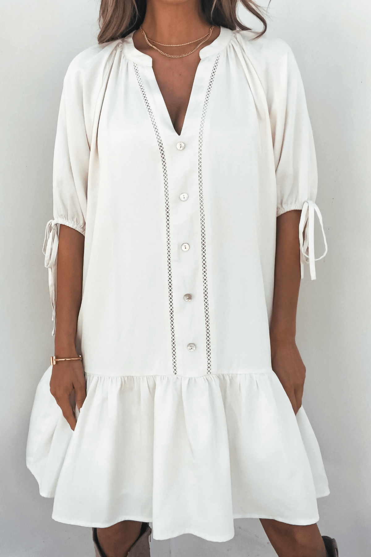 Woman in the Laurie Cream Button Down Mini Dress—flowy, knee-length, with tie sleeves and ruffled hem, against a white background.