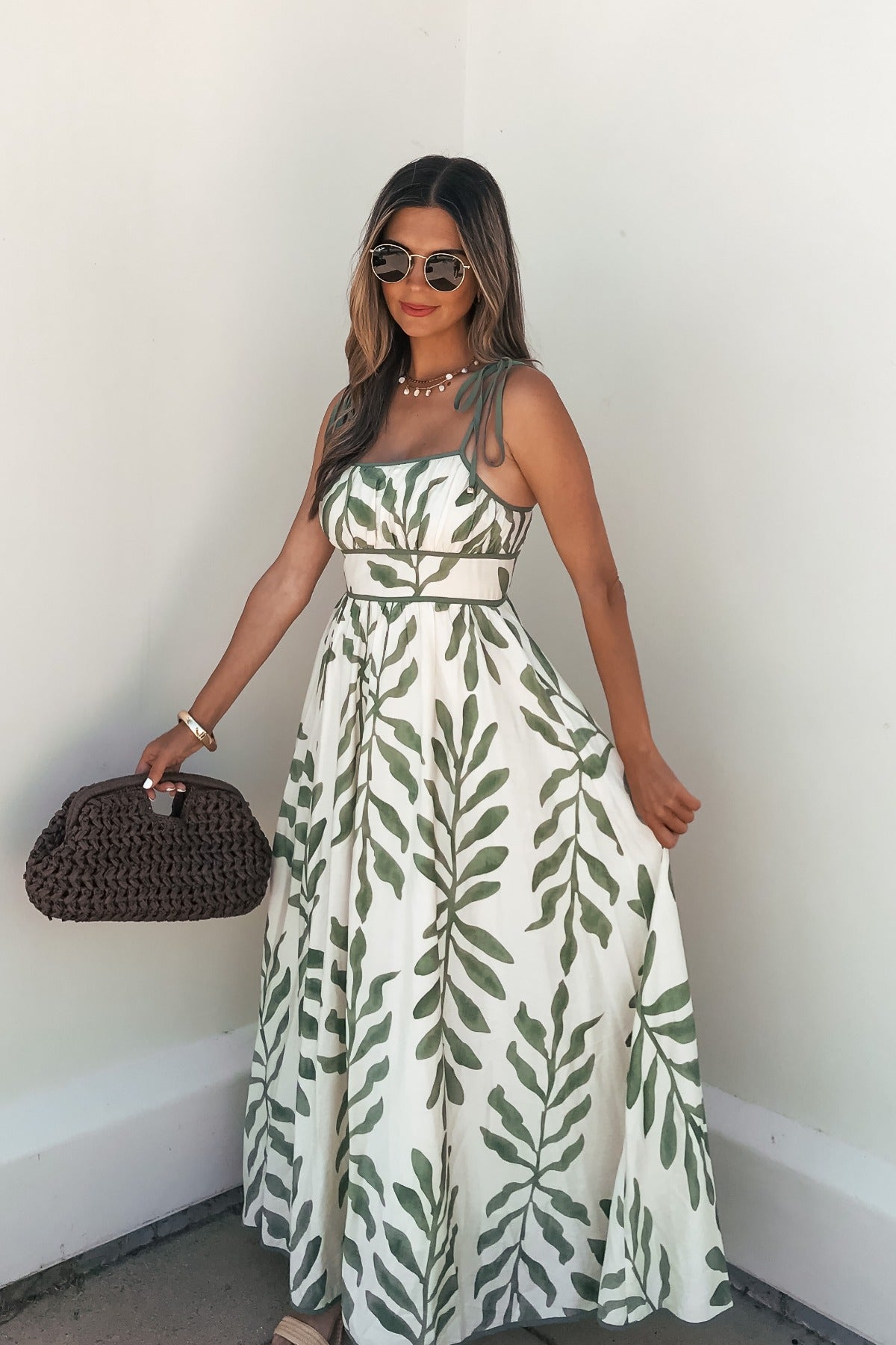 Woman in a Leaf Print Tie Strap Maxi Dress - Green, with sunglasses and a brown woven bag, stands by a white wall.