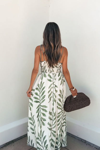 Woman wears Leaf Print Tie Strap Maxi Dress - Green, faces a white wall, holding a brown woven handbag behind her.