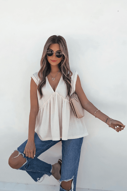 Woman in sunglasses wears the Lena's Carefree Afternoon Cream Babydoll Top, ripped jeans, and a beige bag against a white wall.