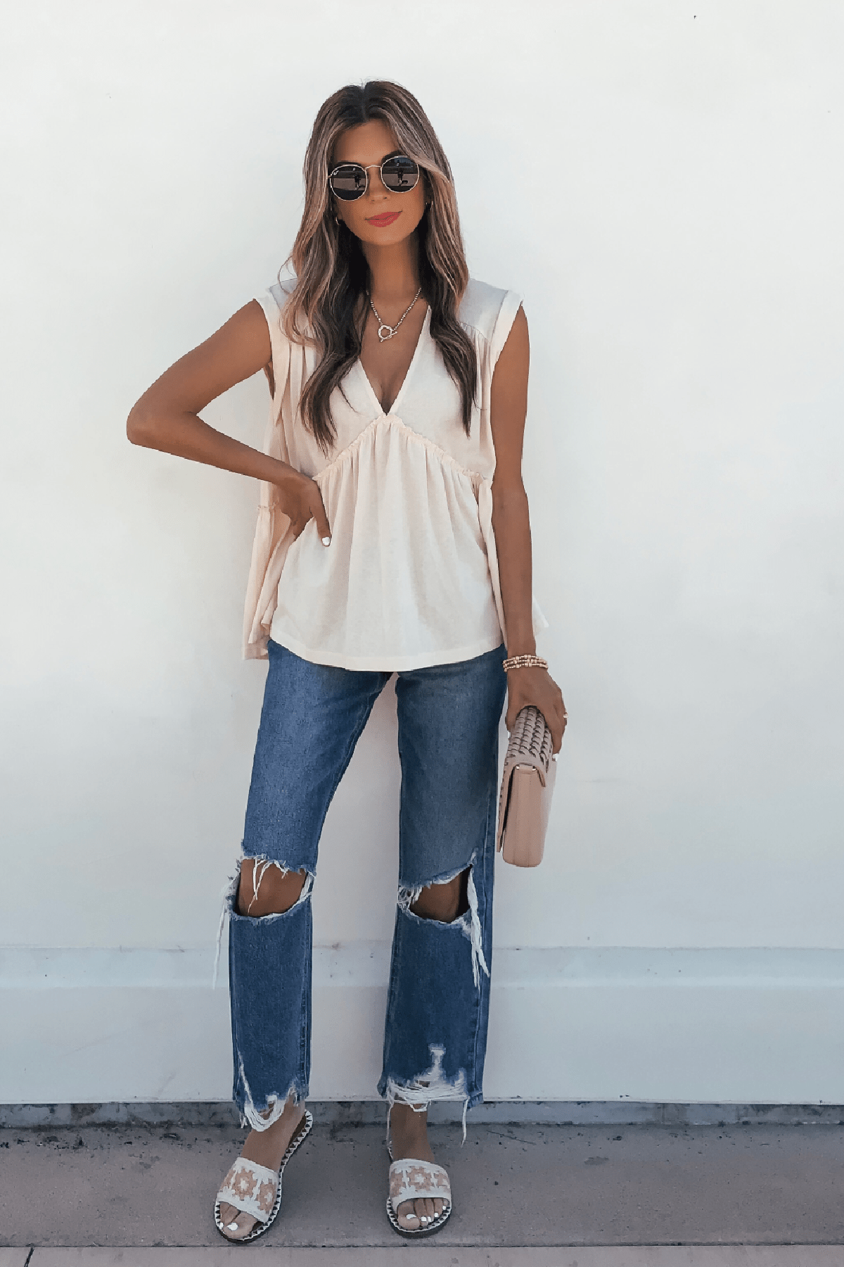 Woman in sunglasses wears Lena's Carefree Afternoon Cream Babydoll Top, ripped jeans, sandals, and a beige clutch by a white wall.